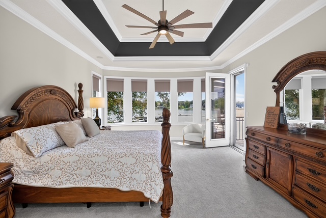 The primary suite with a carved king bed, tray ceiling, bay of lake-facing windows and a private balcony door