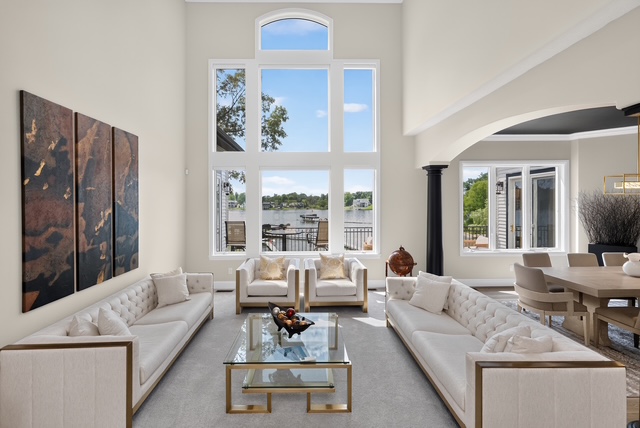 The two-storey great room with tufted sofas, glass coffee table and a wall of windows framing the lake