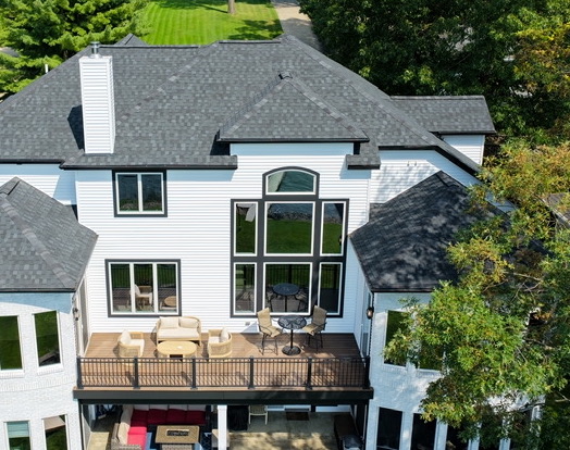 Daytime aerial view of the lake-facing rear elevation of the estate, with the furnished upper deck, covered lower patio and open lawn